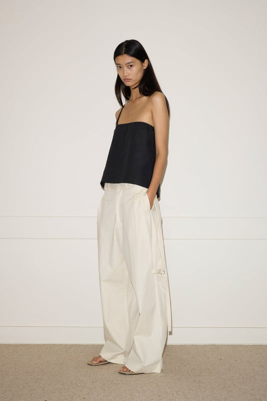 A woman with long, straight black hair stands against a white wall, wearing 0’s off-white Tab Pant—loose, high-waisted wide-leg trousers—with her hands in the pockets, paired with a black strapless top and open-toed sandals.