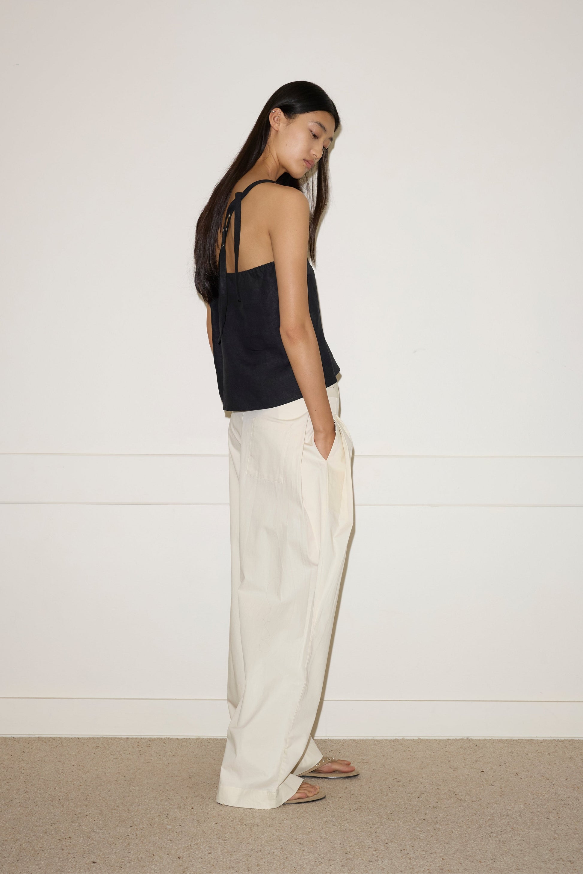 A person with long dark hair stands sideways, wearing the Deiji Studios Button Strap Top - Black with loose white linen wide-leg pants and sandals, one hand in their pocket against a plain light background.