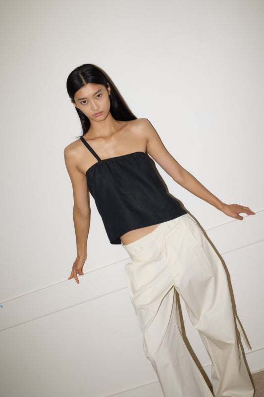 A woman with long dark hair wears the 0 Tab Pant in Off White, paired with a black asymmetrical tank top. She stands against a plain white wall, looking at the camera with a neutral expression.