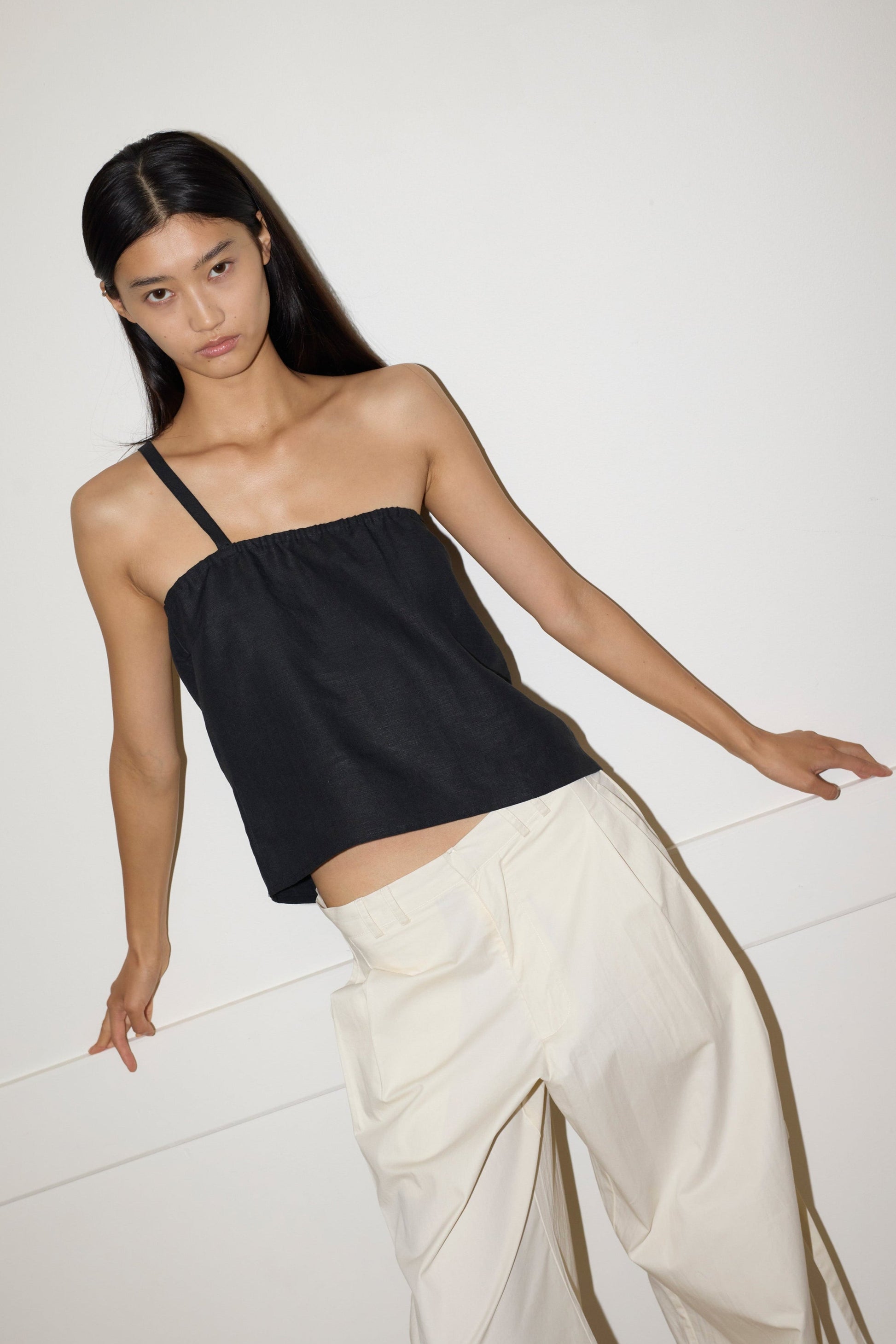 A person with long dark hair wears the Button Strap Top in black and loose cream-colored pants, leaning casually against a white wall.