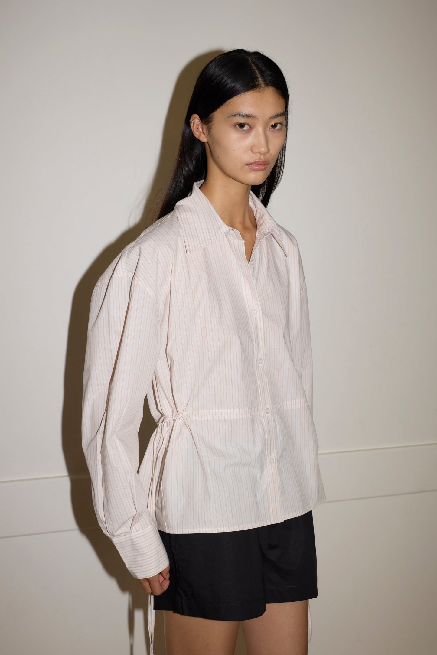 A woman with long, straight black hair wears the Deiji Studios Room Shirt - Dusty Stripe, a loose, light-colored striped organic cotton shirt with side ties, paired with black shorts. She stands against a plain light wall with a neutral expression.