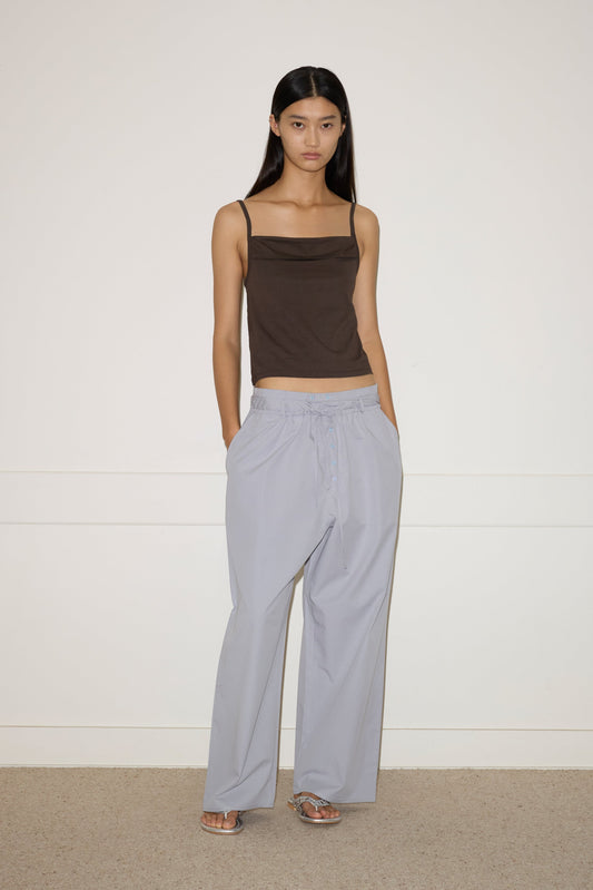 A woman with long straight hair and a neutral expression stands by a white wall, wearing the 0 Two Band Pant in Cornflower, a dark brown sleeveless top, and silver open-toe sandals.