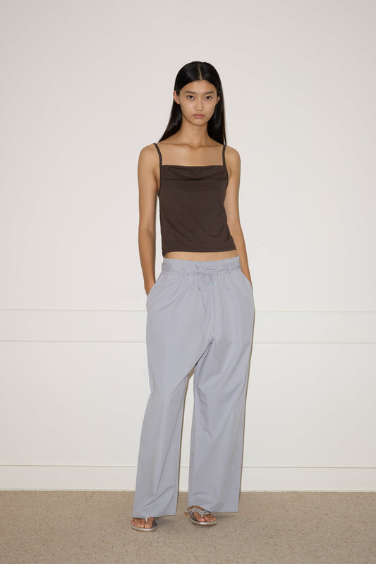A woman with long dark hair stands against a plain wall, wearing the 0 Simple Tank in Caribou, loose light gray pants, and silver open-toe heels. She has a neutral expression and her hands are in her pockets.