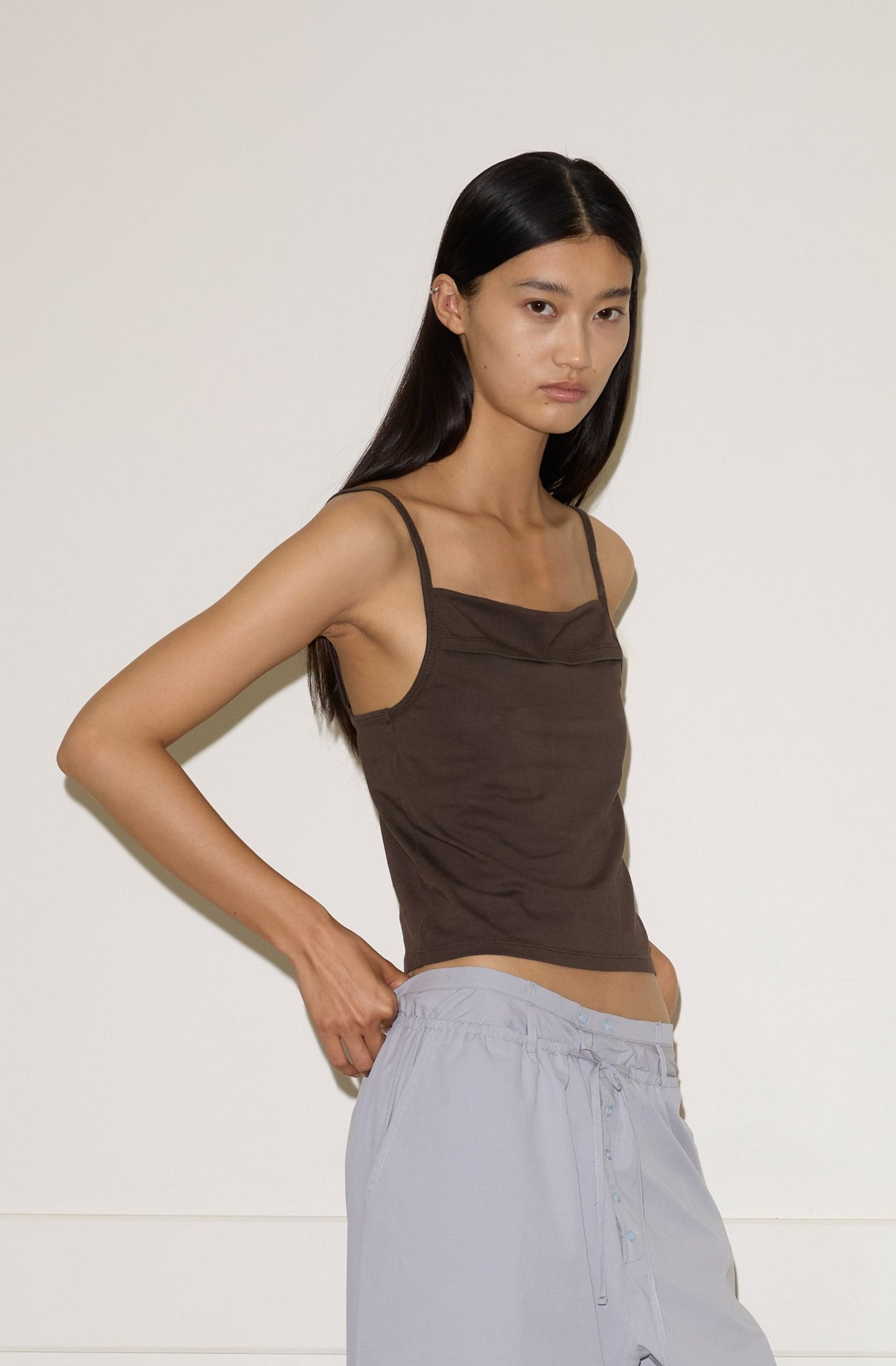 A woman with long dark hair stands against a plain wall, wearing the 0 Simple Tank - Caribou, loose light gray pants, and open-toe sandals. She poses with one hand on her hip, looking confidently at the camera.