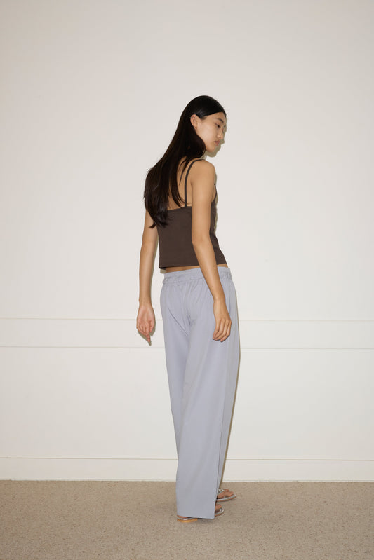 A woman with long dark hair, wearing the 0 Simple Tank - Caribou and light gray loose pants, stands in a minimalist room with a white wall and beige carpet, looking over her shoulder.