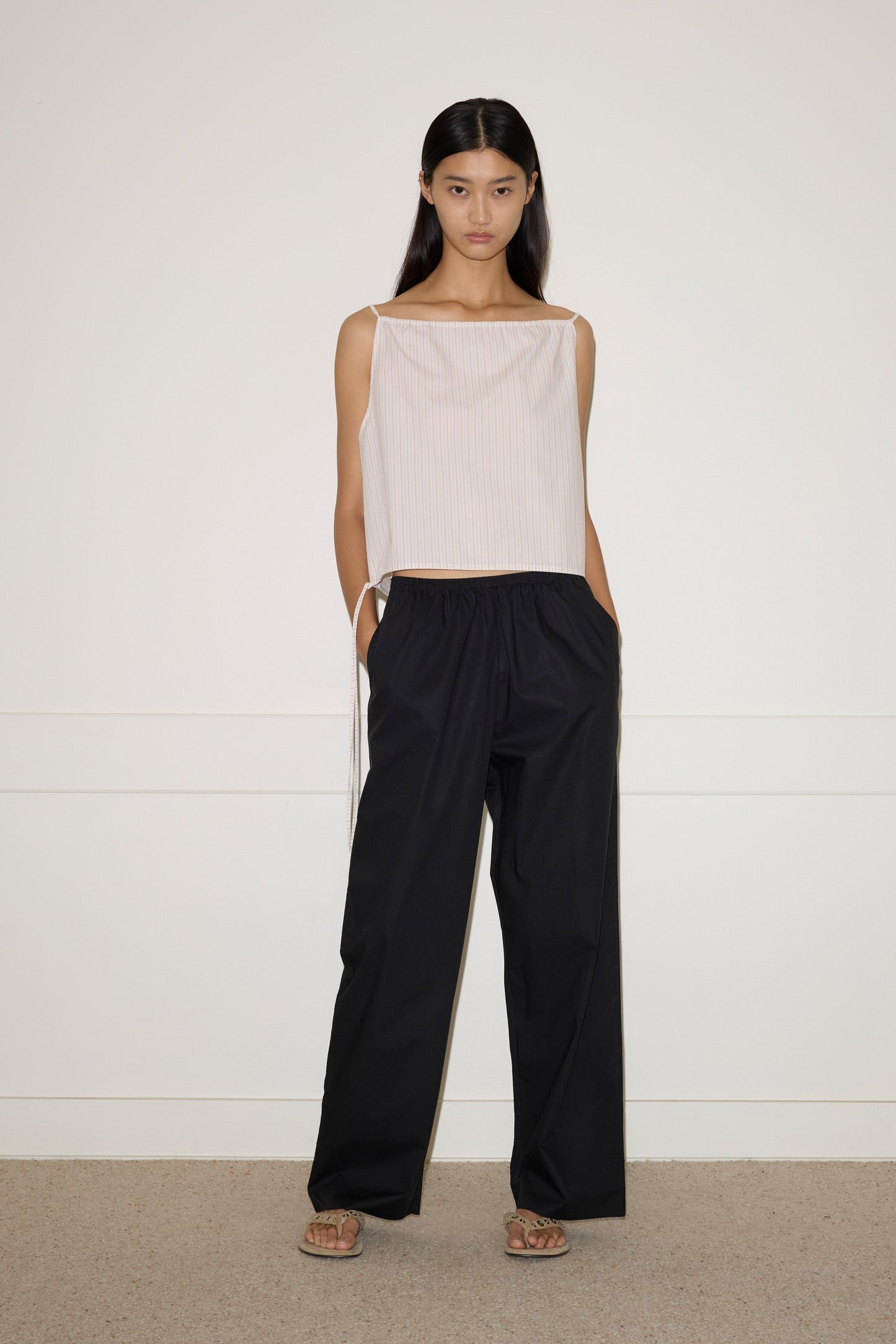 A woman with long dark hair stands indoors against a plain white wall, wearing the sleeveless, loose beige Simple Top and wide-leg black pants. She completes the look with sandals and has her hands in her pockets.