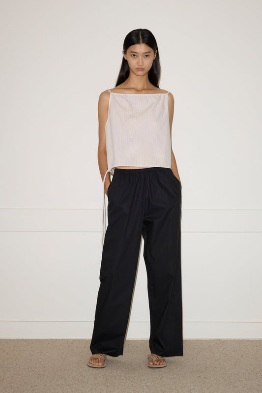 A woman with long dark hair stands indoors against a plain white wall, wearing the sleeveless, loose beige Simple Top and wide-leg black pants. She completes the look with sandals and has her hands in her pockets.