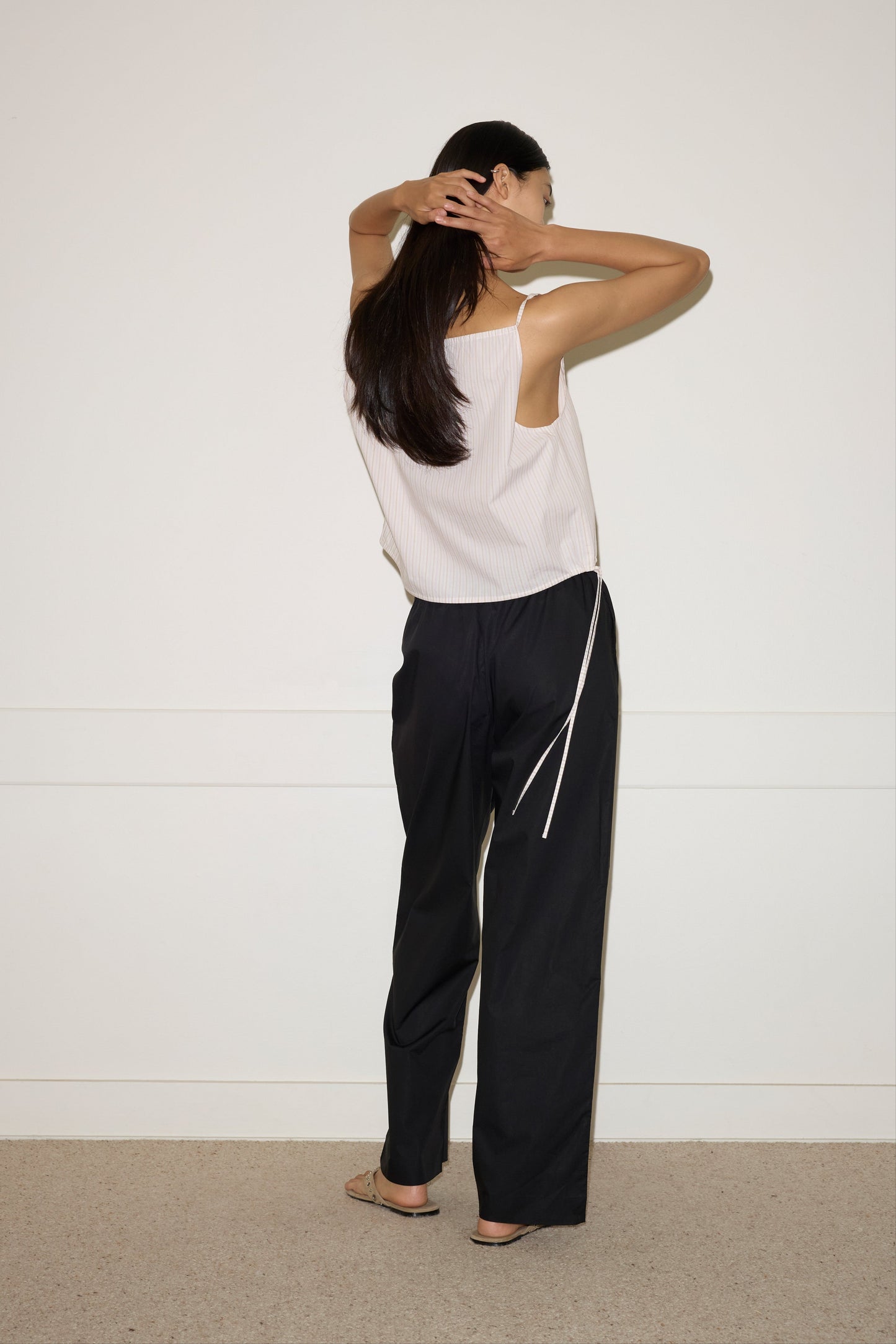 A woman with long dark hair stands facing a white wall, wearing the Simple Top - Dusty Stripe by Deiji Studios and loose black pants, her arms raised with hands touching her head.