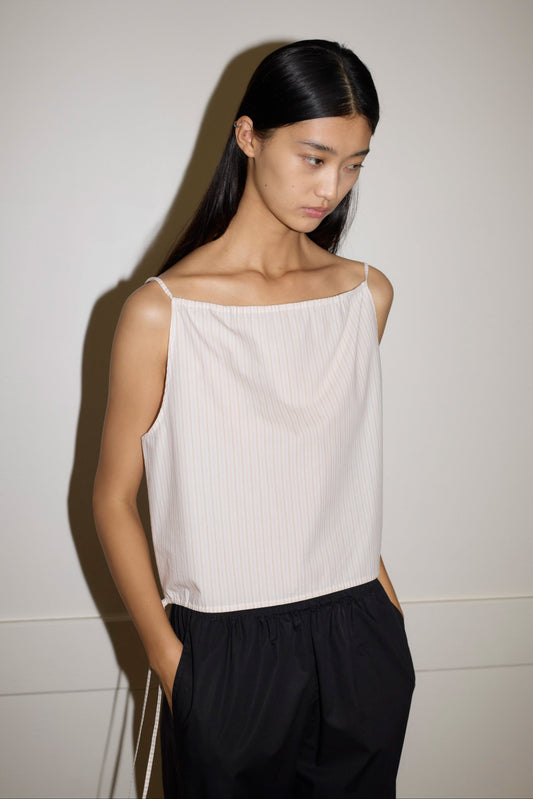 A woman with long, straight black hair stands against a light wall, hands in her pockets and looking down thoughtfully, wearing the Simple Top—a sleeveless, light-colored striped piece—paired with loose black pants.