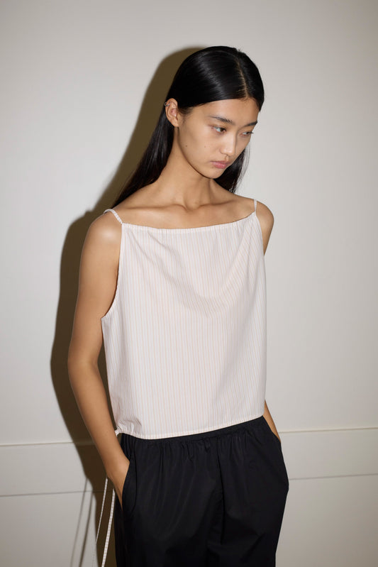 A woman with long, straight black hair stands against a light wall, hands in her pockets and looking down thoughtfully, wearing the Simple Top—a sleeveless, light-colored striped piece—paired with loose black pants.