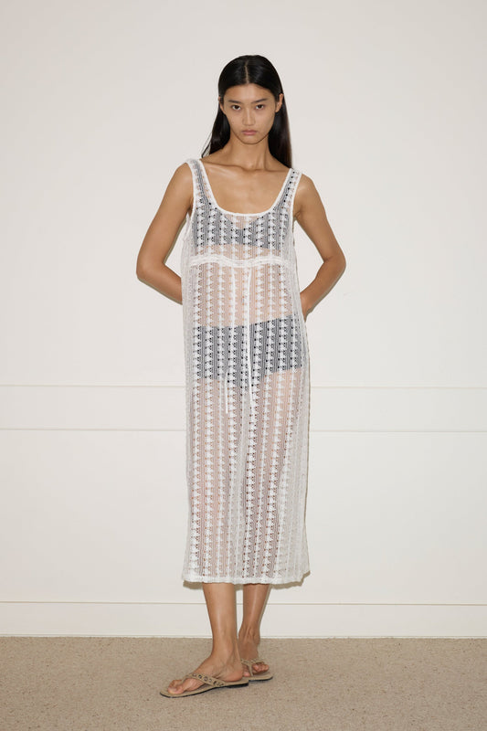 A woman with long, straight hair poses against a plain background wearing the Thread Dress - Off White by 0—a sleeveless, sheer crochet lace dress layered over a black bikini with flat sandals and her arms relaxed behind her back.