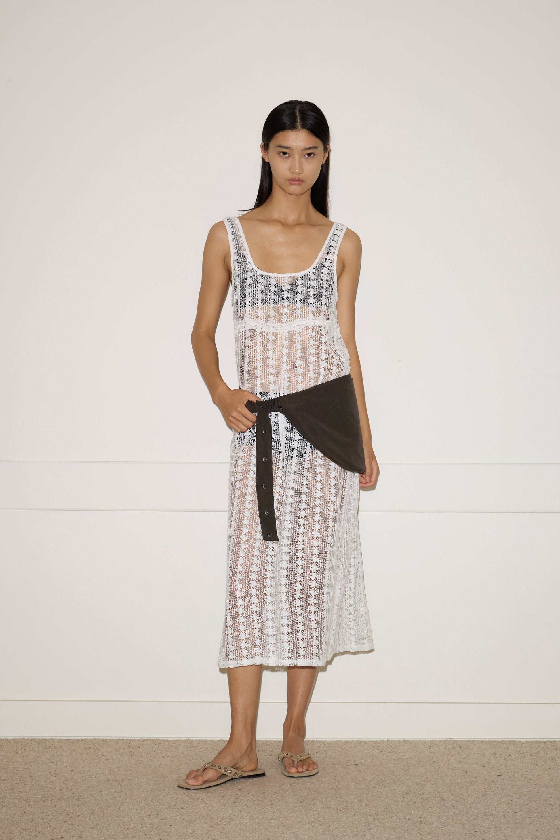 A woman with straight, long dark hair and a neutral expression stands against a plain wall, wearing the 0 Thread Dress - Off White over a black bikini, paired with a dark shirt at her waist and beige sandals.