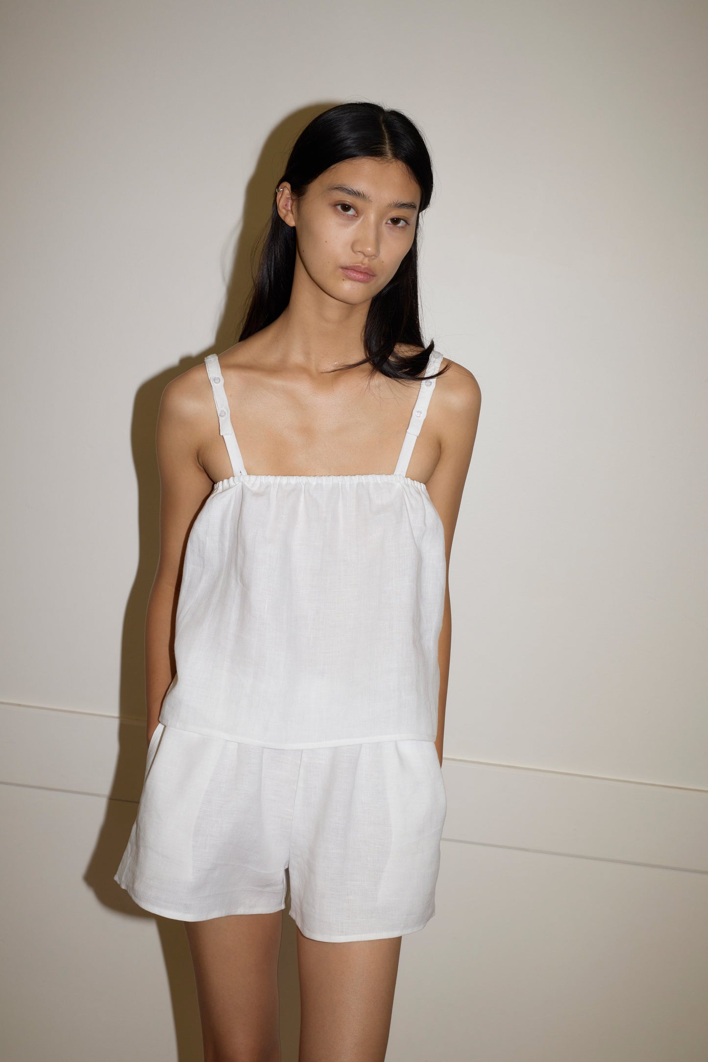 A woman with long, straight black hair stands against a plain wall, wearing the Button Linen Set - White. The sleeveless outfit features thin straps and matching shorts. She has a neutral expression and her hands are in her pockets.