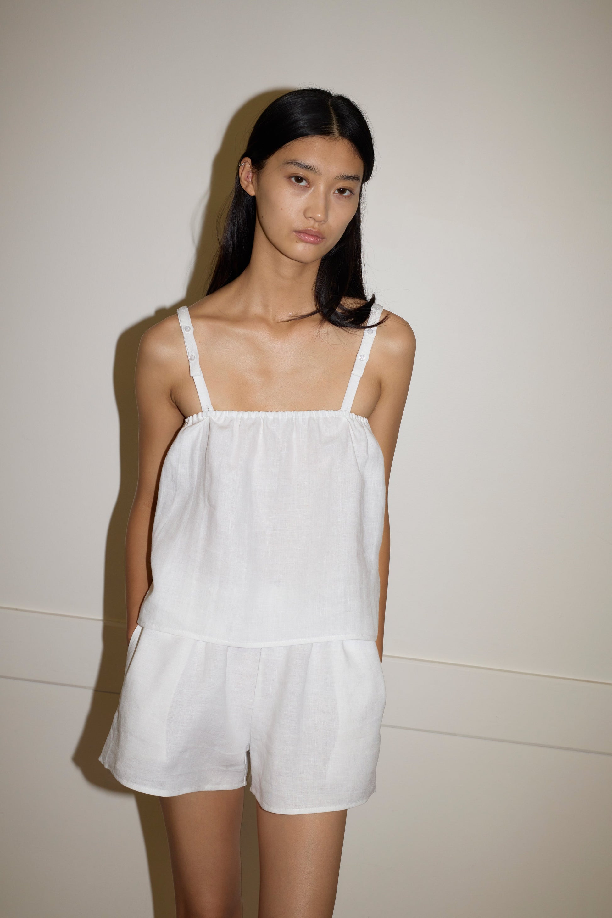 A woman with long, straight black hair stands against a plain wall, wearing the Button Linen Set - White. The sleeveless outfit features thin straps and matching shorts. She has a neutral expression and her hands are in her pockets.