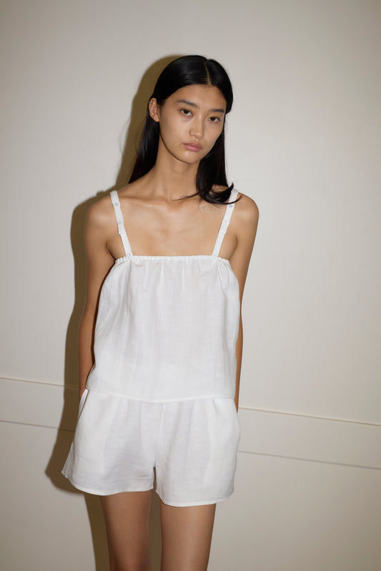 A woman with long, straight black hair stands against a plain wall, wearing the Button Linen Set - White. The sleeveless outfit features thin straps and matching shorts. She has a neutral expression and her hands are in her pockets.