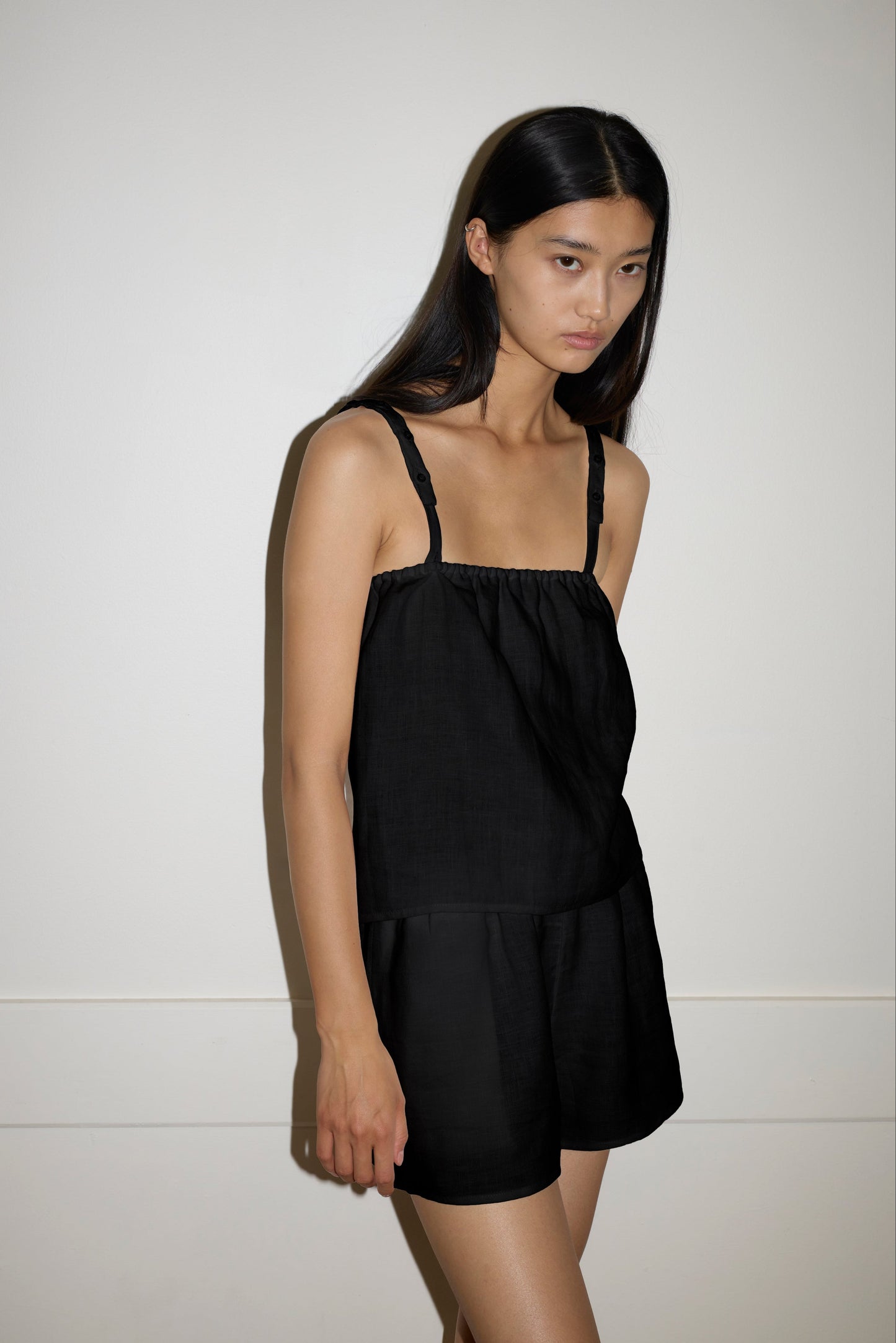 A woman with long dark hair stands against a plain white wall, wearing the sleeveless black Button Linen Set by Deiji Studios. She faces slightly to the side and gazes ahead with a calm, neutral expression.