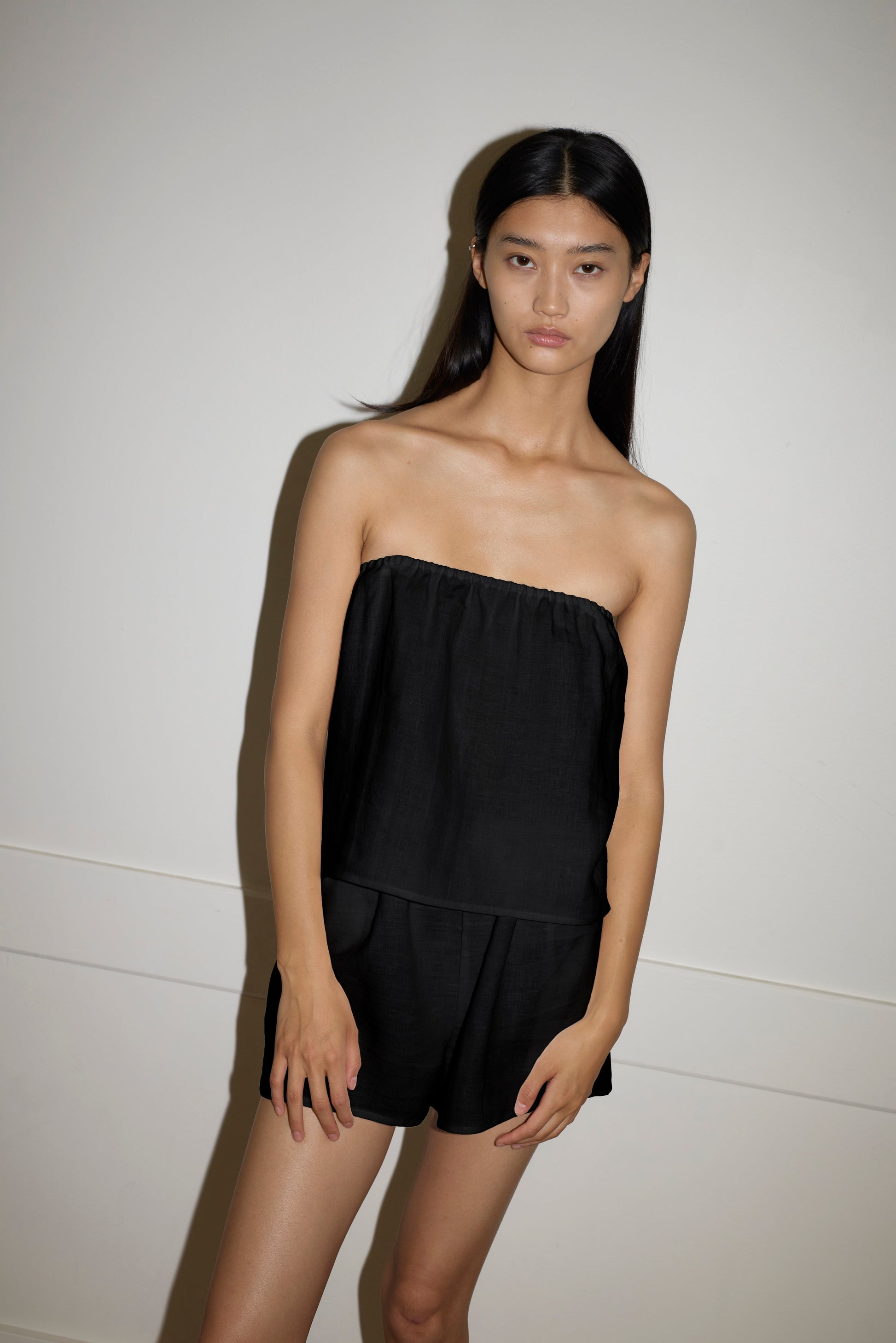 A person with long, straight black hair stands against an off-white wall, wearing a sleeveless, strapless Button Linen Set - Black by Deiji Studios. Their expression is neutral and their posture relaxed.
