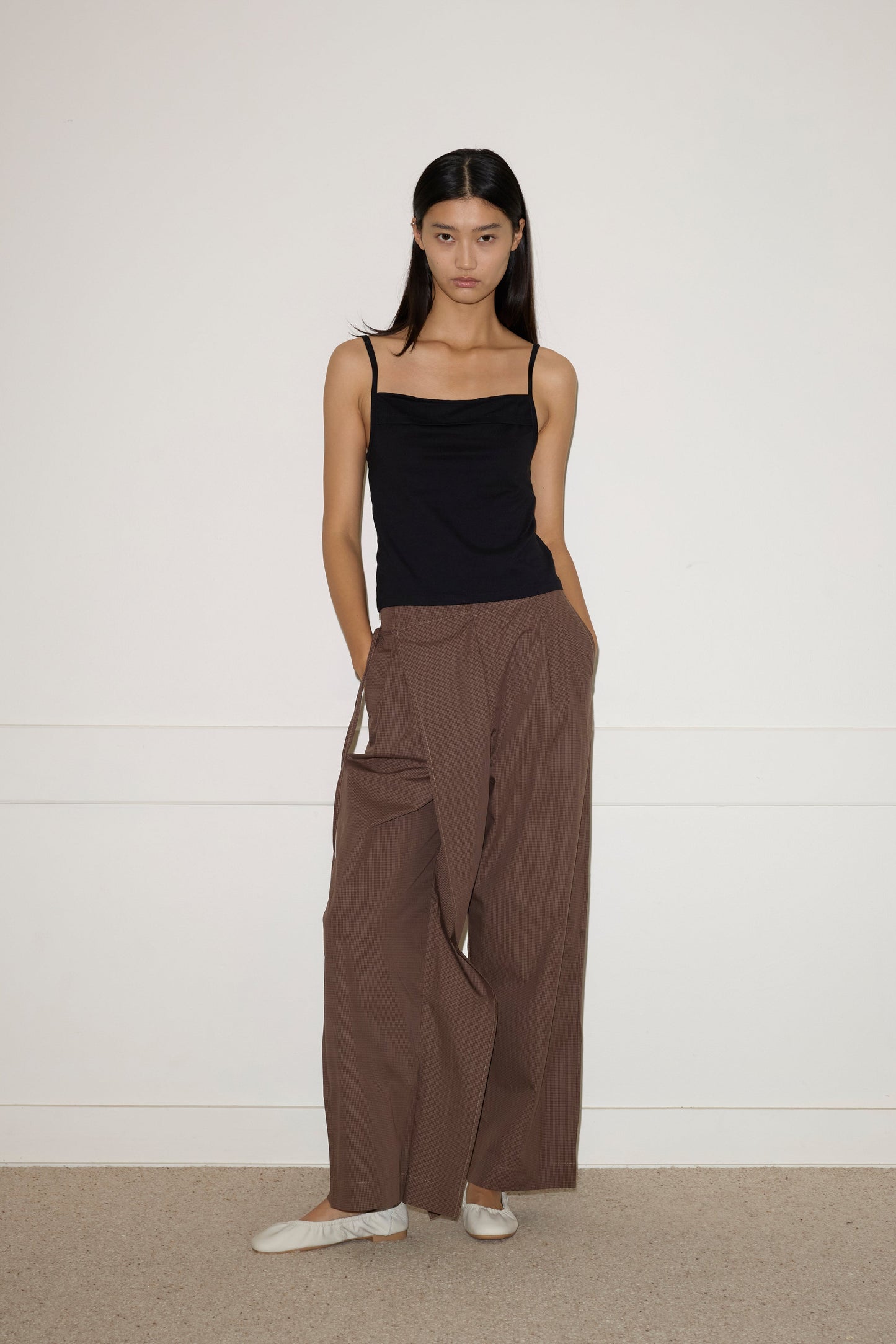 A woman stands against a white wall, wearing the 0 Simple Tank in black, loose brown draped pants, and white shoes. Her hands are in her pockets as she looks directly at the camera.