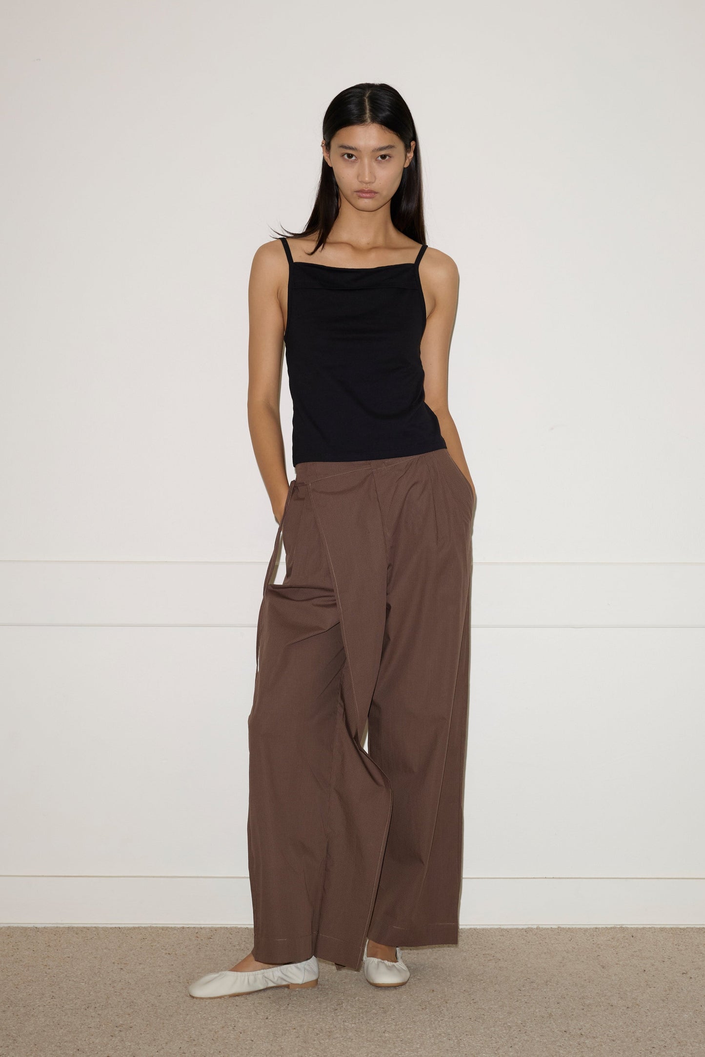 A woman stands against a white wall wearing Double Layer Pant in brown, a black sleeveless top, and white ballet flats. Her hands are in her pockets and she has long, straight dark hair.