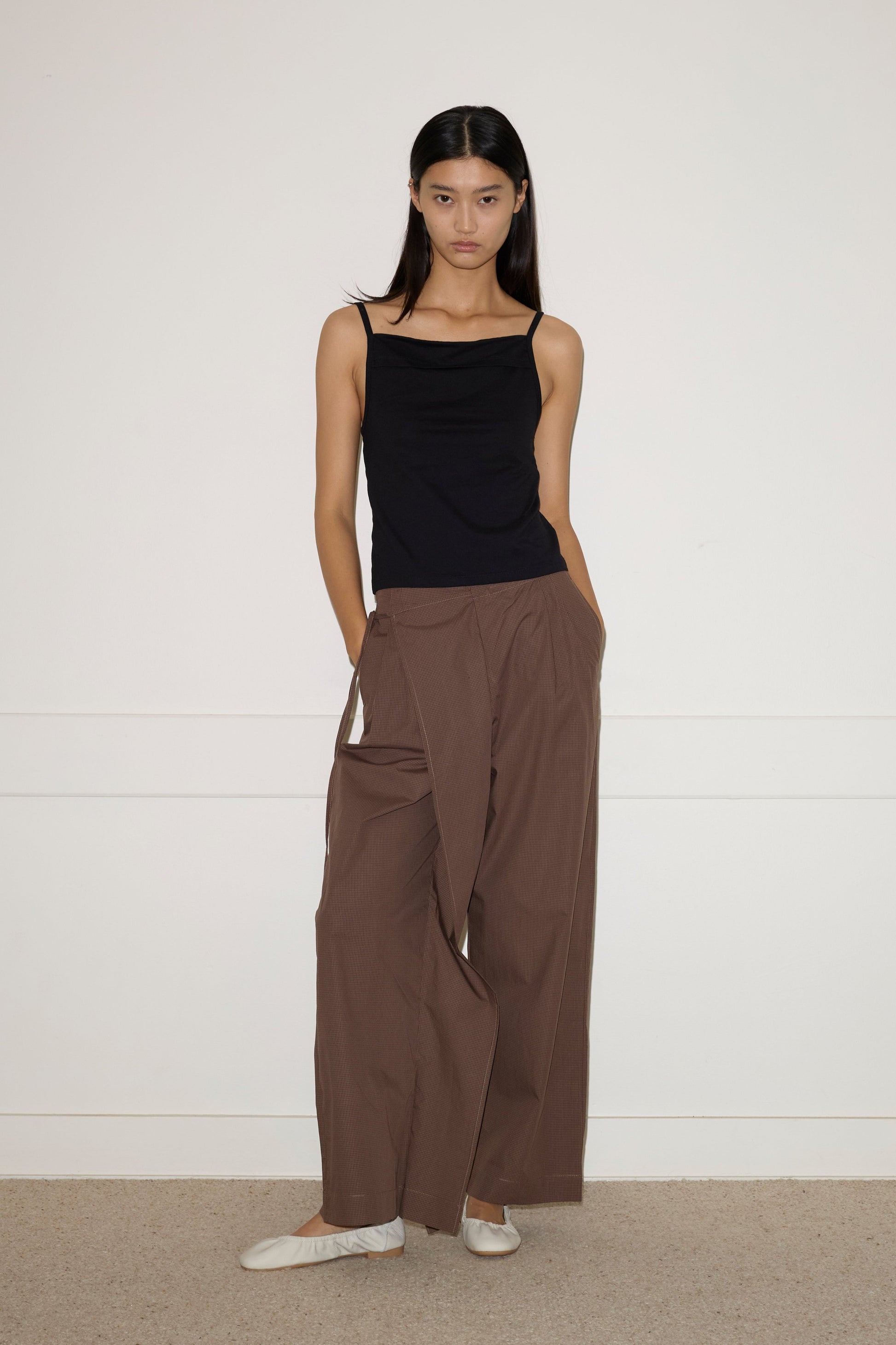A woman stands against a white wall wearing Double Layer Pant in brown, a black sleeveless top, and white ballet flats. Her hands are in her pockets and she has long, straight dark hair.