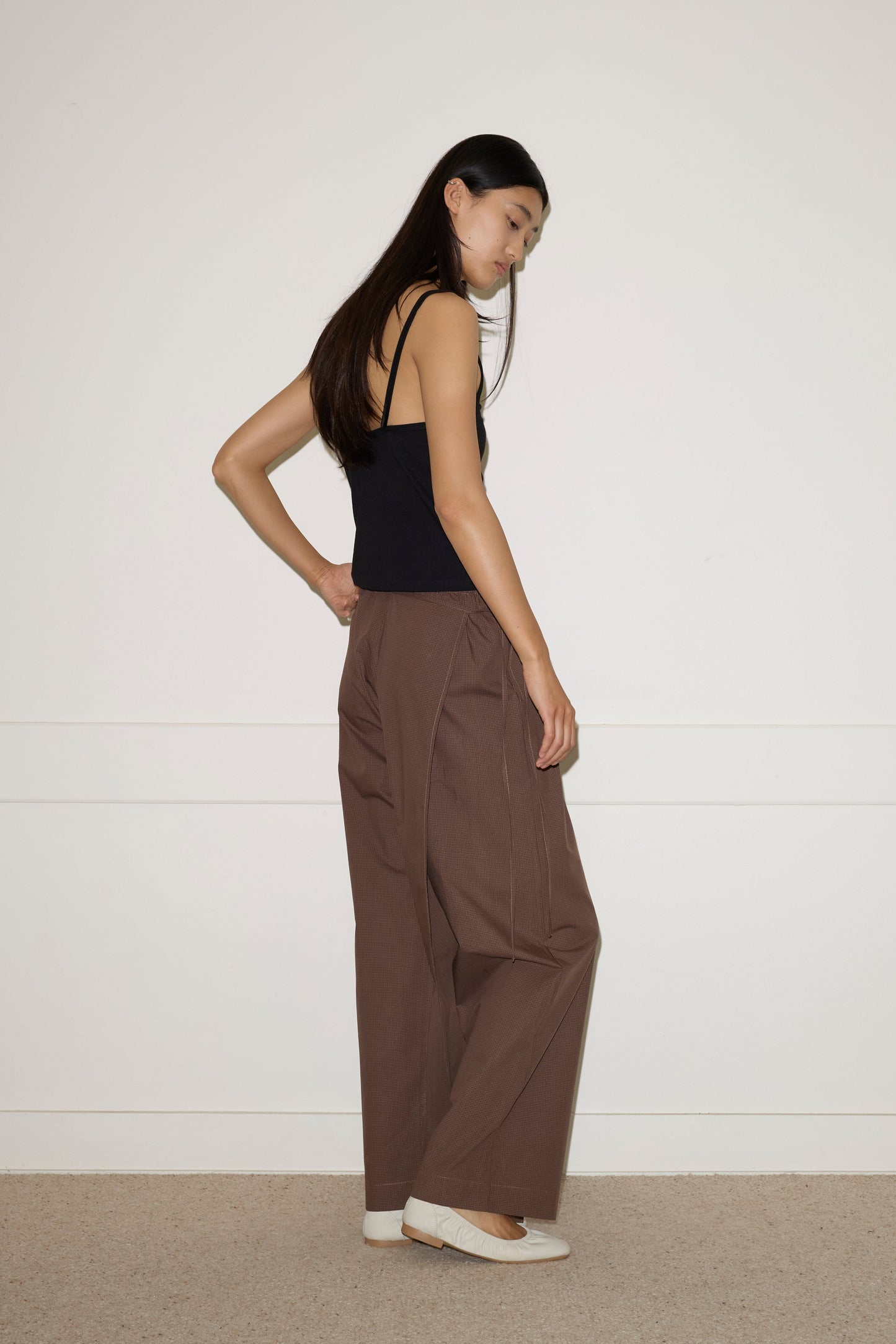 A woman with long dark hair stands sideways, wearing a black sleeveless top and the Deiji Studios Double Layer Pant - Carob Check in loose organic cotton, paired with white flats, against a plain white wall and beige carpeted floor.