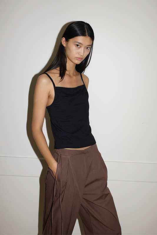 A person with long dark hair stands against a plain light wall, wearing the Simple Tank - Black by Deiji Studios and loose brown pants, one hand in their pocket and a neutral expression.