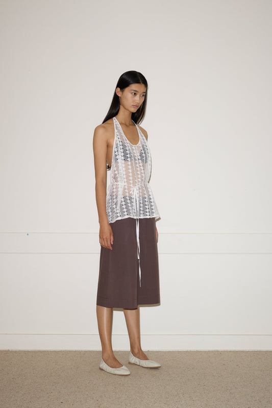 A woman with long dark hair and a neutral expression stands indoors against a plain wall, wearing the 0 Thread Wrap Top - Off White over a nude underlayer, paired with a knee-length brown skirt and light-colored flats.