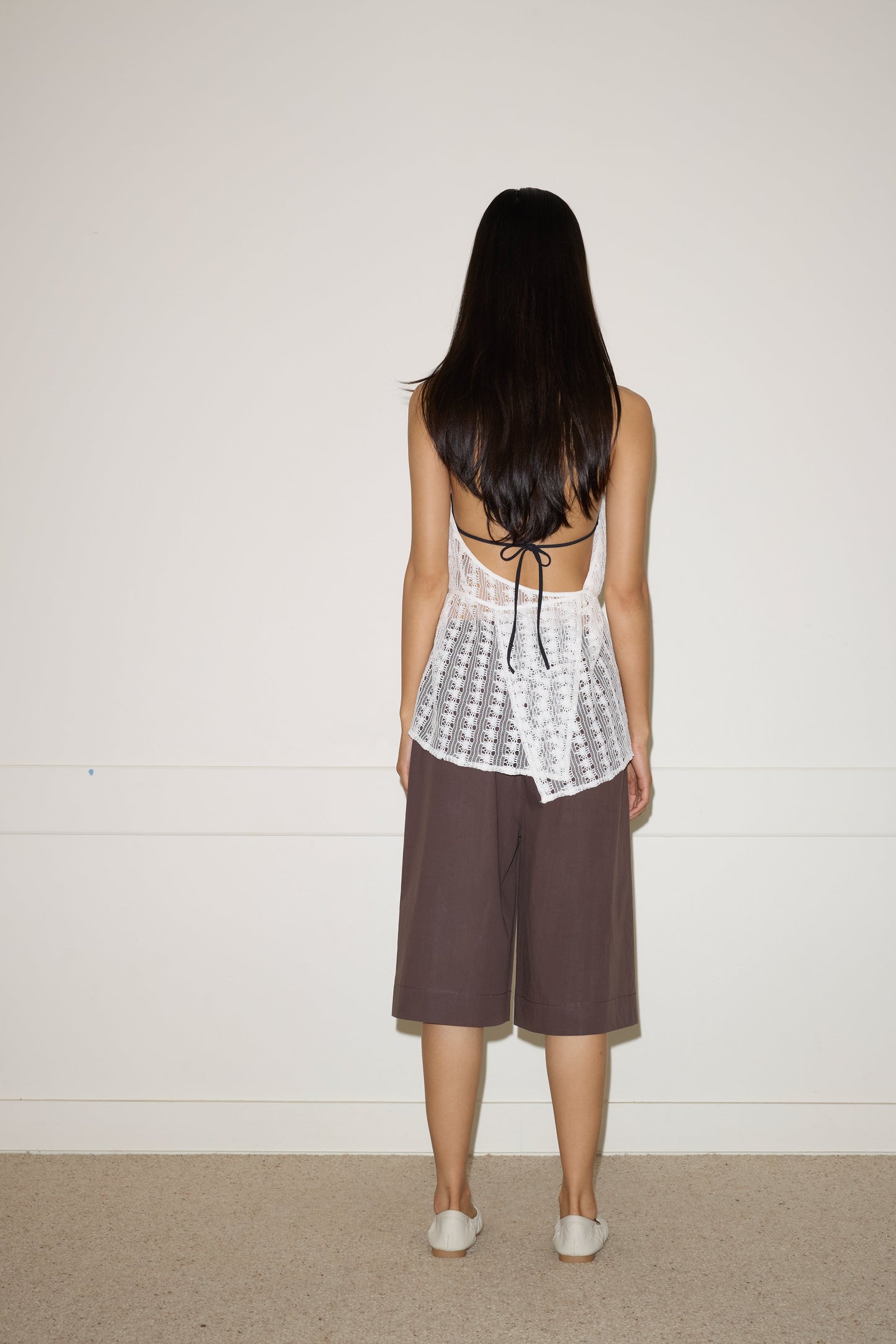 A woman with long dark hair faces a plain white wall, wearing the 0 Thread Wrap Top - Off White paired with loose brown knee-length shorts and white shoes.