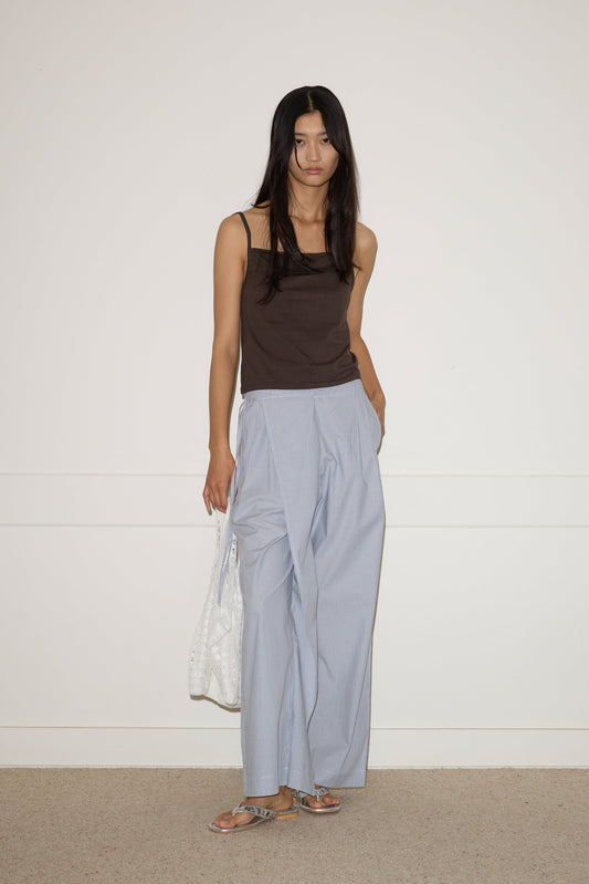 A woman with long dark hair stands against a white wall, wearing a sleeveless dark brown top, 0’s Double Layer Pant in Sky Stripe, silver sandals, and holding a white patterned tote bag.