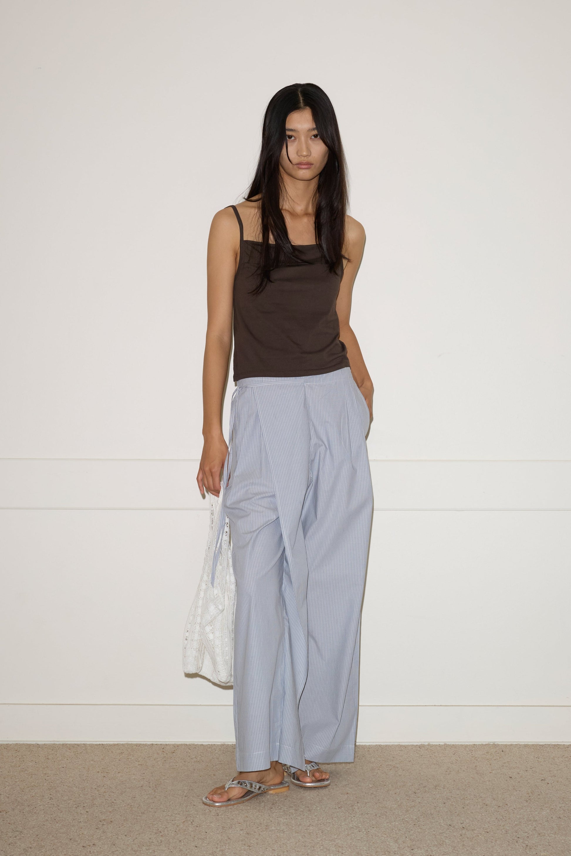 A woman with long dark hair stands against a white wall, wearing a sleeveless dark brown top, 0’s Double Layer Pant in Sky Stripe, silver sandals, and holding a white patterned tote bag.