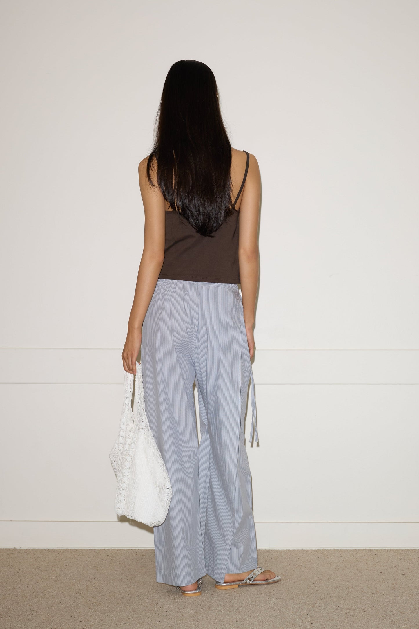 A woman with long dark hair faces a blank white wall, wearing a brown sleeveless top, sandals, holding a large white tote bag, and light blue Double Layer Pant - Sky Stripe by 0.