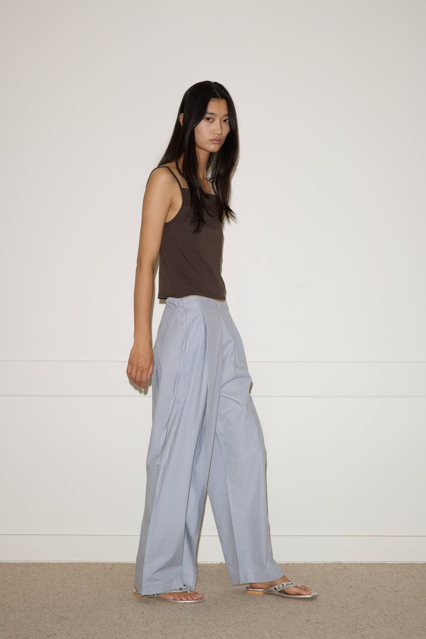 A person with long dark hair, wearing a brown sleeveless top and 0 Double Layer Pant - Sky Stripe, stands against a plain off-white wall and beige carpeted floor.