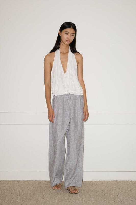A woman with long black hair wears a sleeveless white halter top and Double Waist Linen Pant in Haze Blue, standing against a plain white wall and beige carpeted floor.