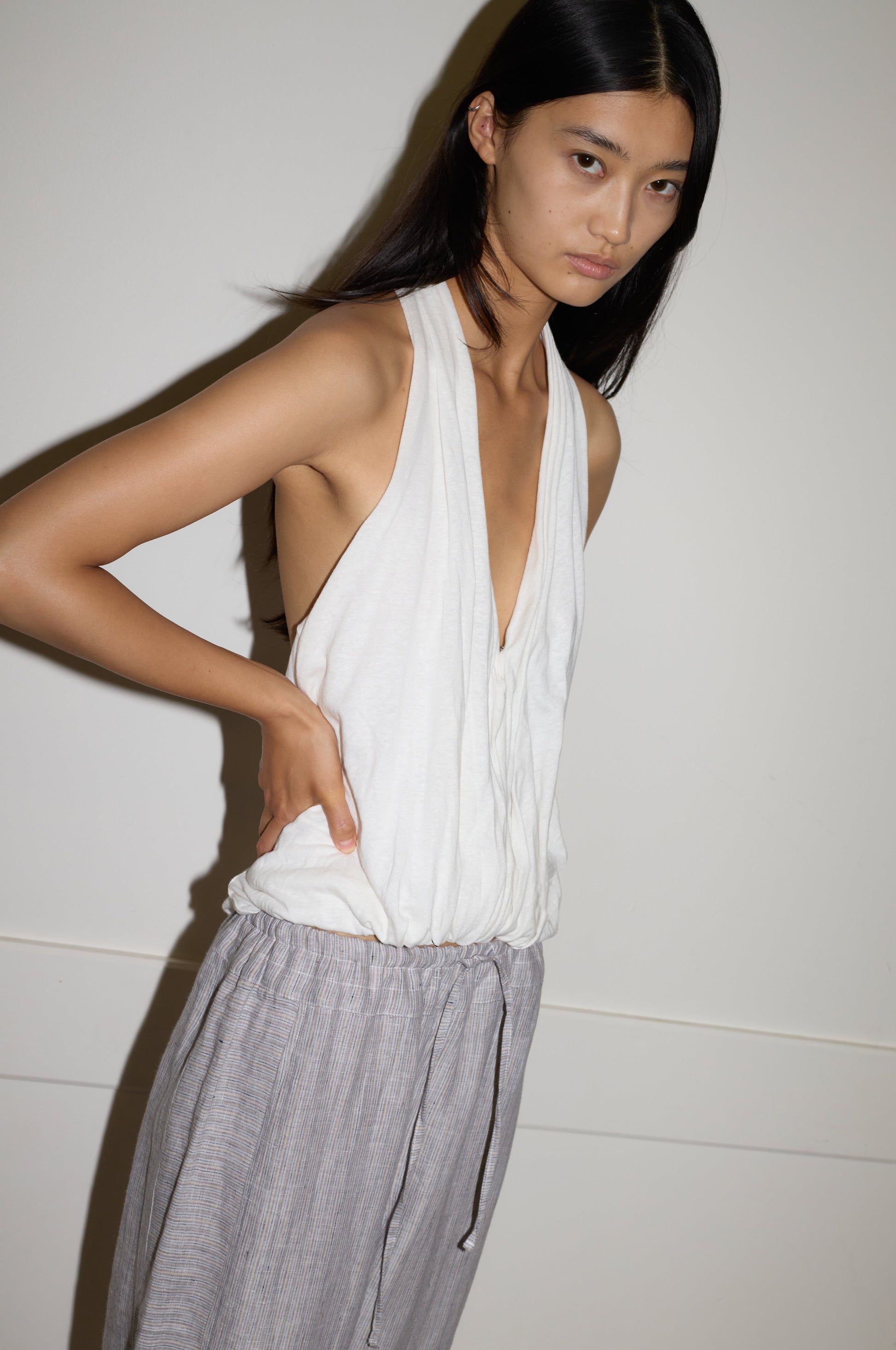 Model wears the Jersey Top - White, a draped deep-V sleeveless design by Deiji Studios, tucked into grey striped drawstring trousers and posed against a neutral wall.