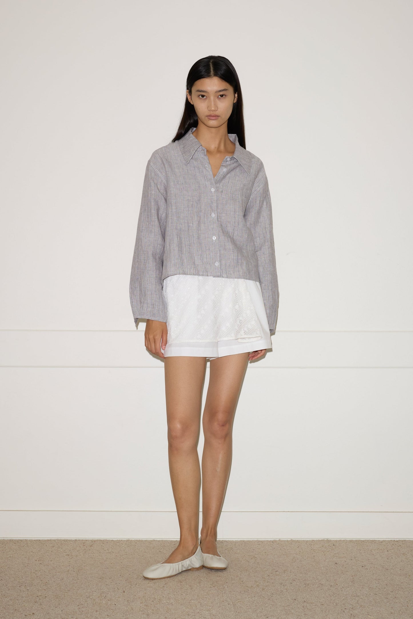 A woman with long dark hair and a neutral expression stands against a plain white wall, wearing the 0 Collar Point Shirt - Haze Blue over a short white skirt and beige flats.