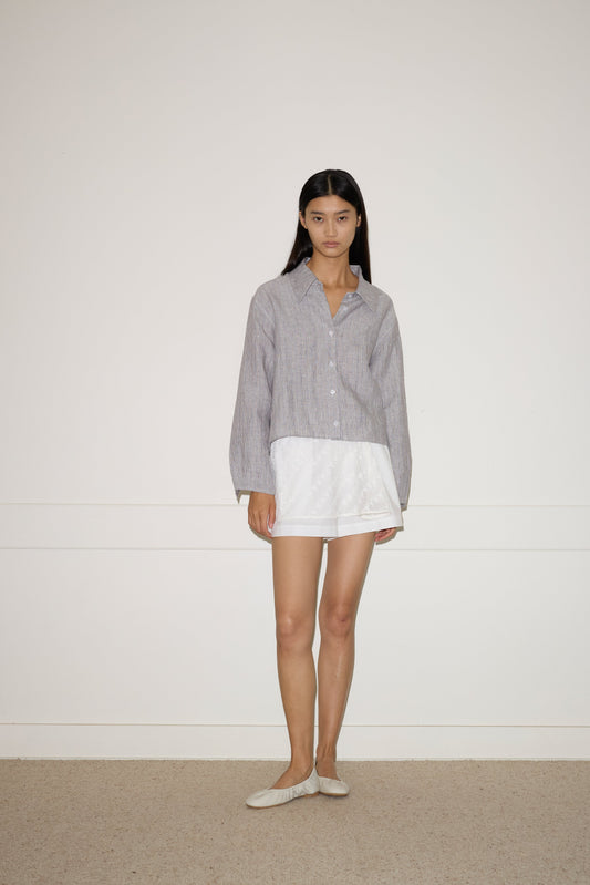 A woman stands indoors against a plain wall, wearing a loose gray Deiji Studios organic cotton button-up shirt over a white skirt and beige flats. Her long straight hair and neutral expression complete the look with the Thread Button Belt in Off White.