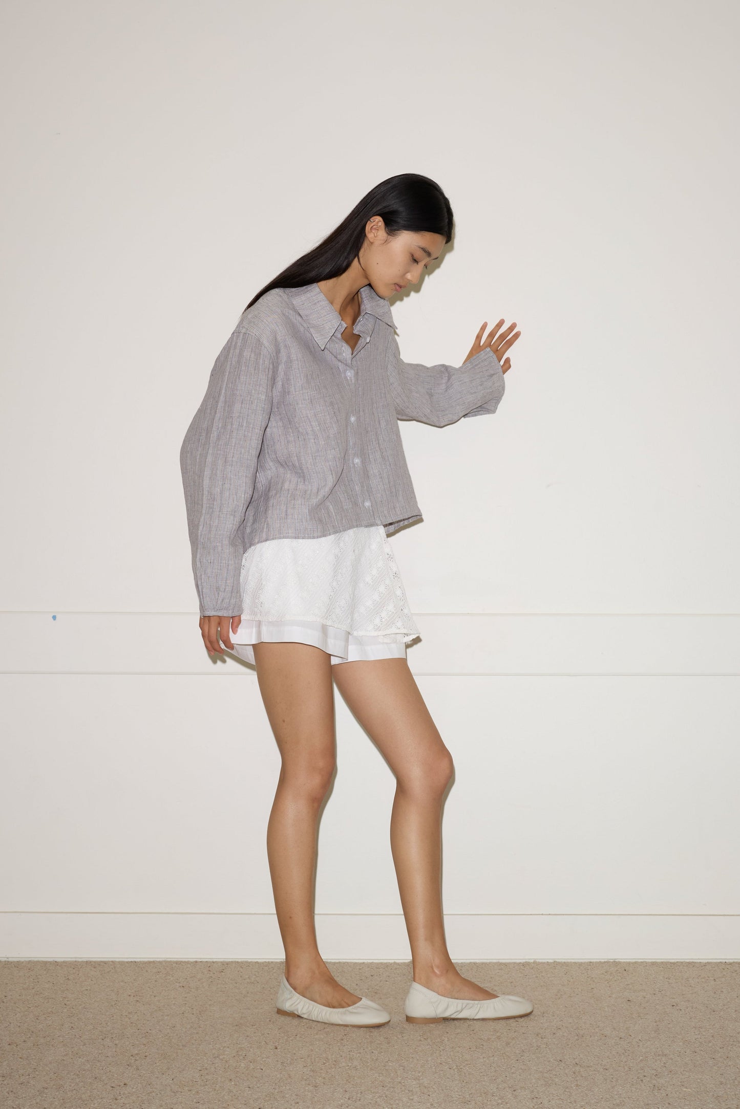 A woman with long dark hair, wearing the 0 Collar Point Shirt in Haze Blue, white shorts, and beige flats stands sideways against a plain white wall, appearing to walk with one arm extended forward.