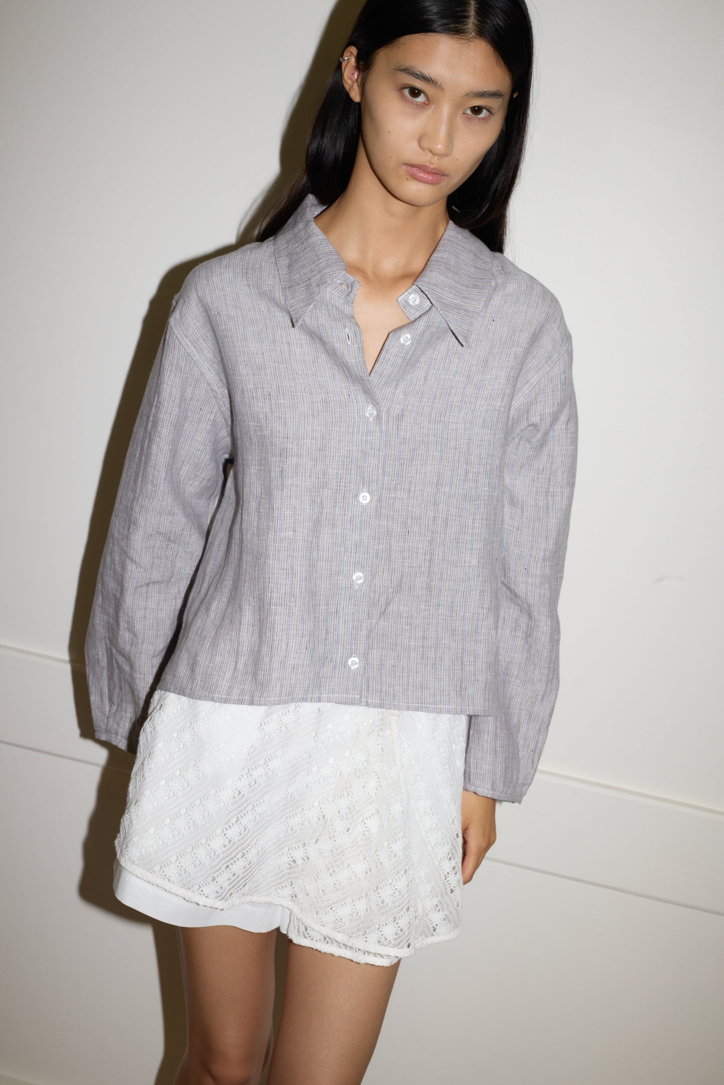 A woman with long black hair wears the 0 Collar Point Shirt in haze blue, paired with a short white lace skirt, standing against a white wall and looking at the camera with a neutral expression.
