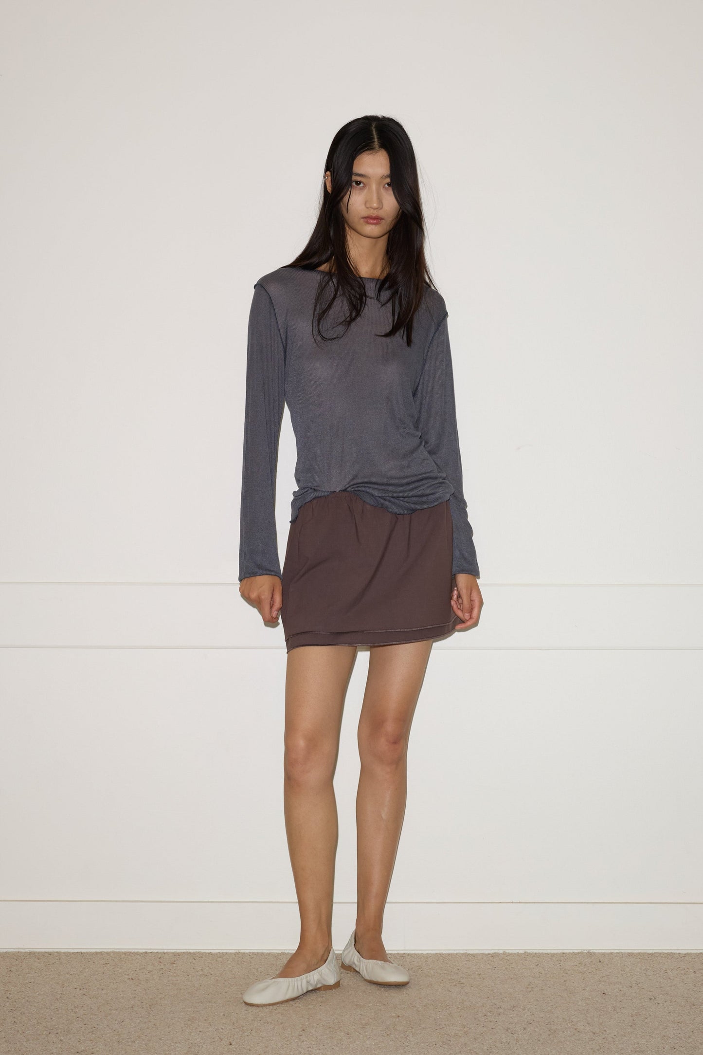 A woman with long dark hair stands against a white wall, wearing the 0 Knit Long Sleeve - Steel, a brown skirt, and white flat shoes. Her posture and expression are relaxed and neutral.