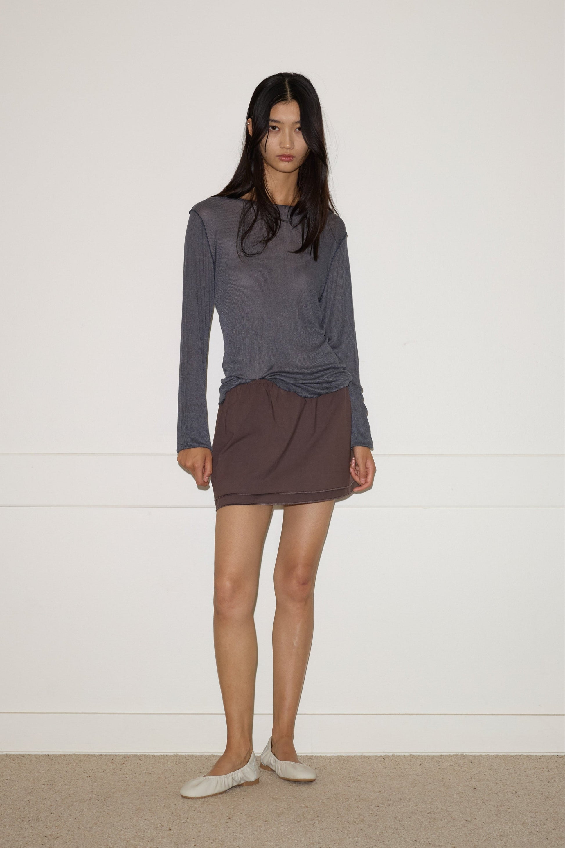A woman with long dark hair stands indoors against a white wall, wearing a loose gray organic cotton top, the 0 Layered Skirt - Chocolate, and white slippers, with a neutral expression and relaxed posture.