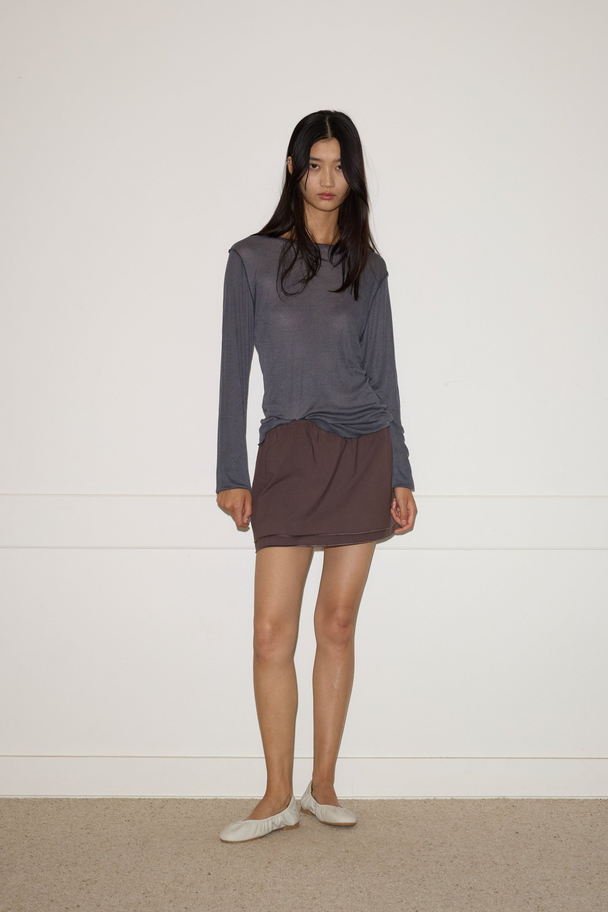 A woman with long dark hair stands indoors against a white wall, wearing a loose gray organic cotton top, the 0 Layered Skirt - Chocolate, and white slippers, with a neutral expression and relaxed posture.