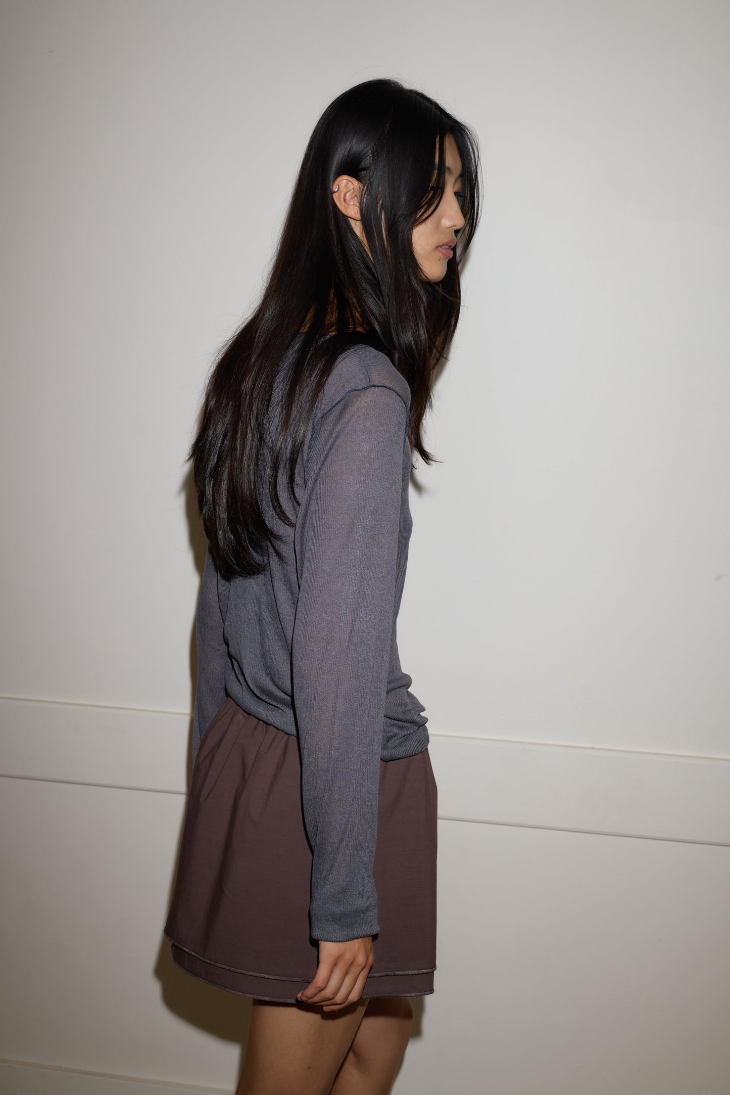 A young woman with long, dark hair stands sideways against a plain white wall, wearing the 0 Knit Long Sleeve - Steel and a brown skirt. She looks down with her arms resting naturally by her sides.