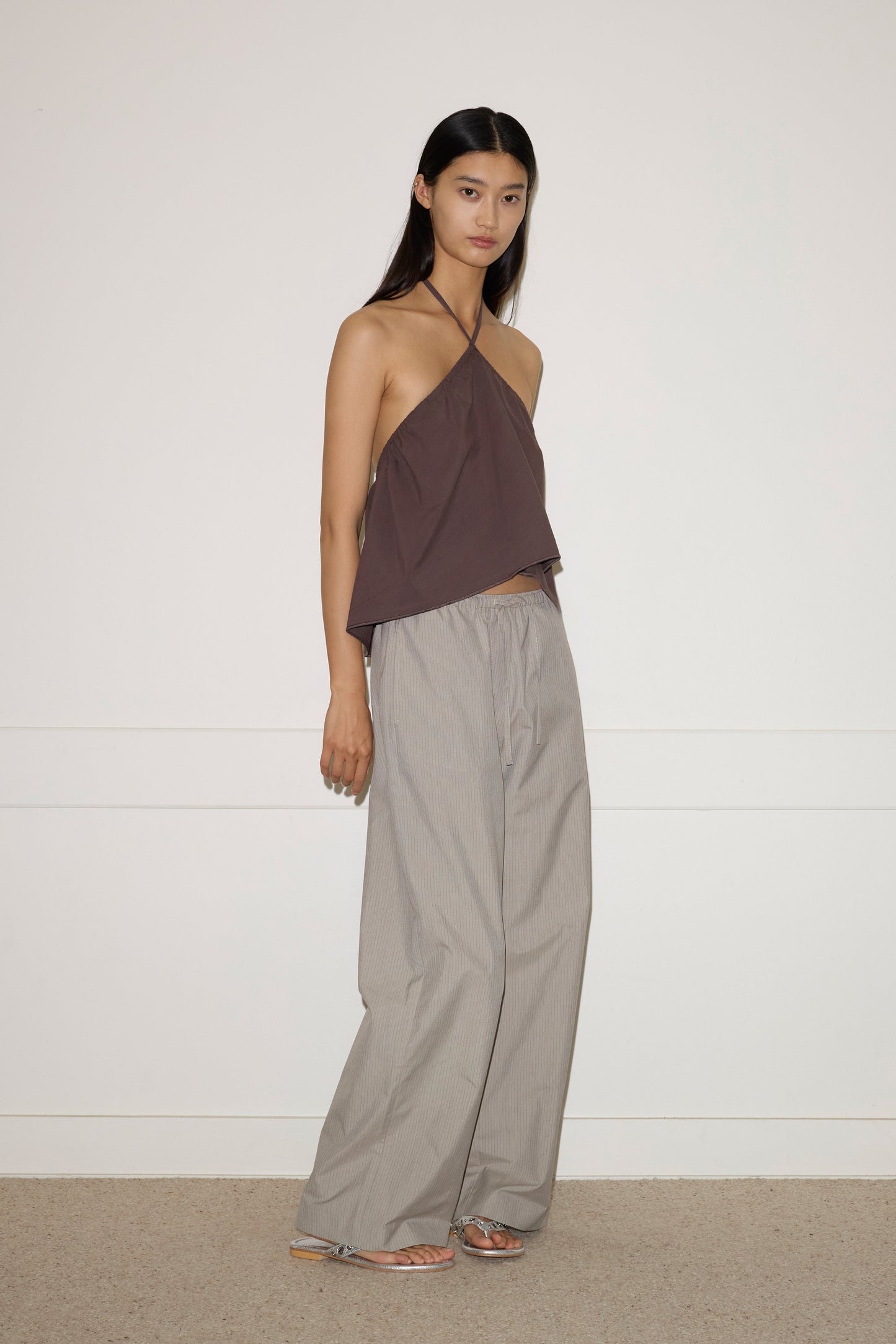 A woman with long, straight black hair and a neutral expression wears the Napkin Top - Chocolate by 0, paired with loose light gray wide-leg pants and metallic sandals against a plain backdrop.
