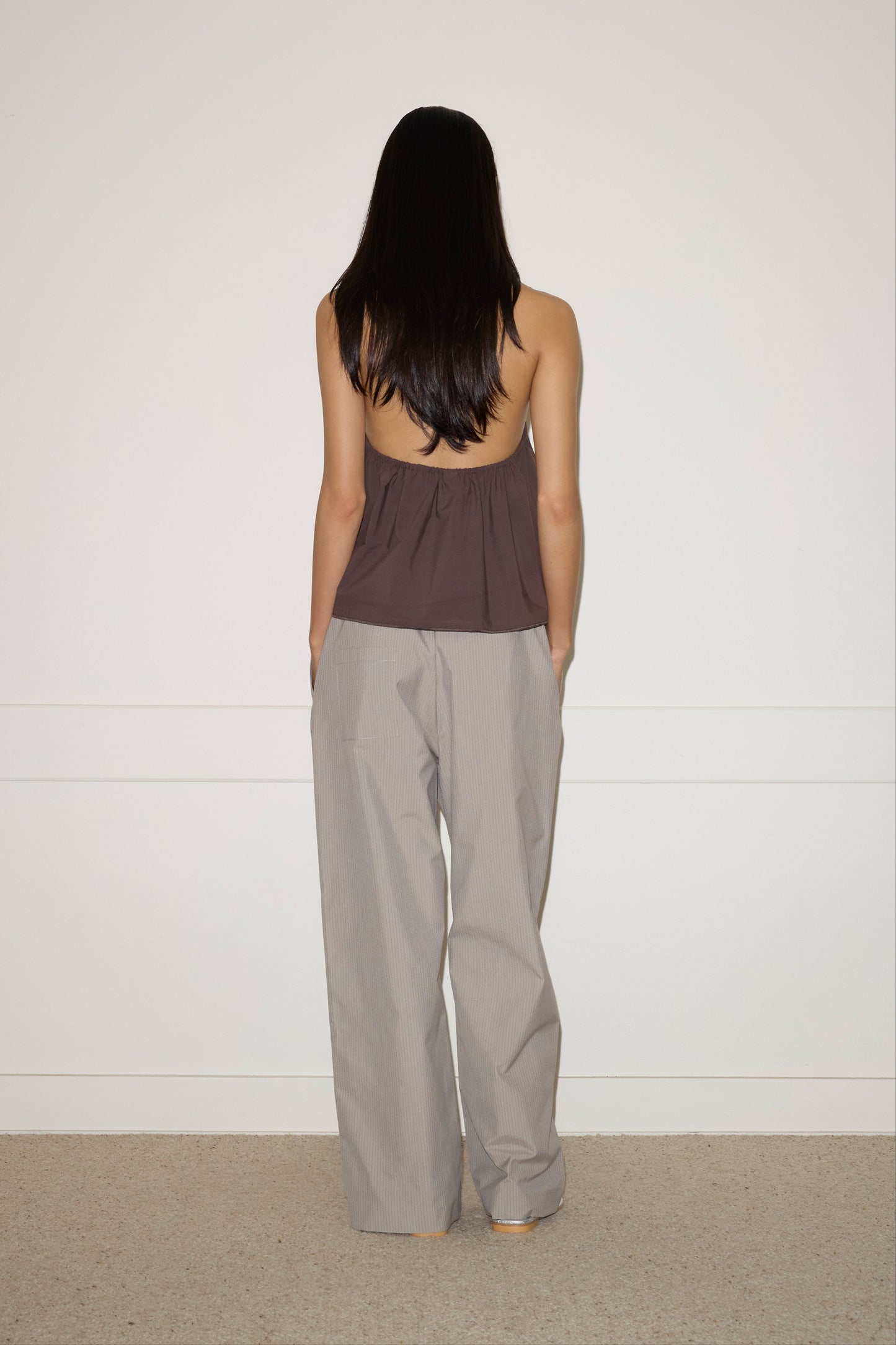 A woman with long dark hair stands facing a white wall, wearing the Chocolate Napkin Top by 0 and loose gray pants, her hands in her pockets.