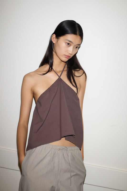 A person with long dark hair wears the 0 Napkin Top in Chocolate and light gray pants, standing against a plain white wall with hands in pockets, gazing slightly downward.