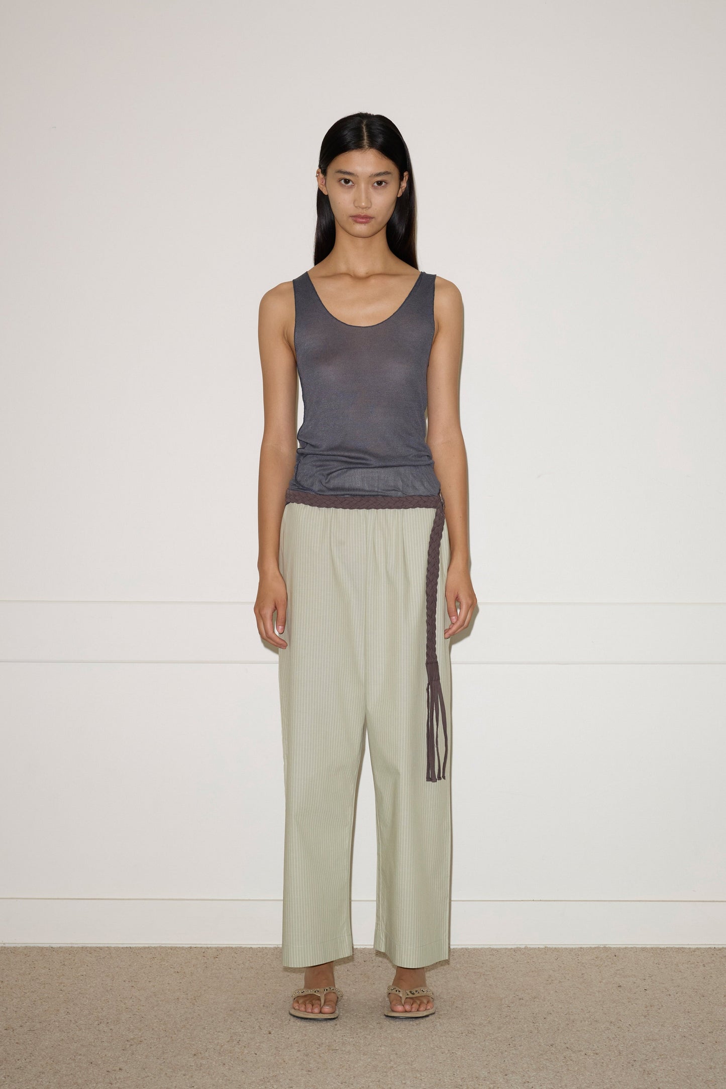 A person stands against a plain white wall, wearing a dark gray sleeveless Deiji Studios top, loose light green organic cotton pants with the 0 Plaited Belt in Chocolate, and sandals. Their hair is long and straight; their expression is neutral.
