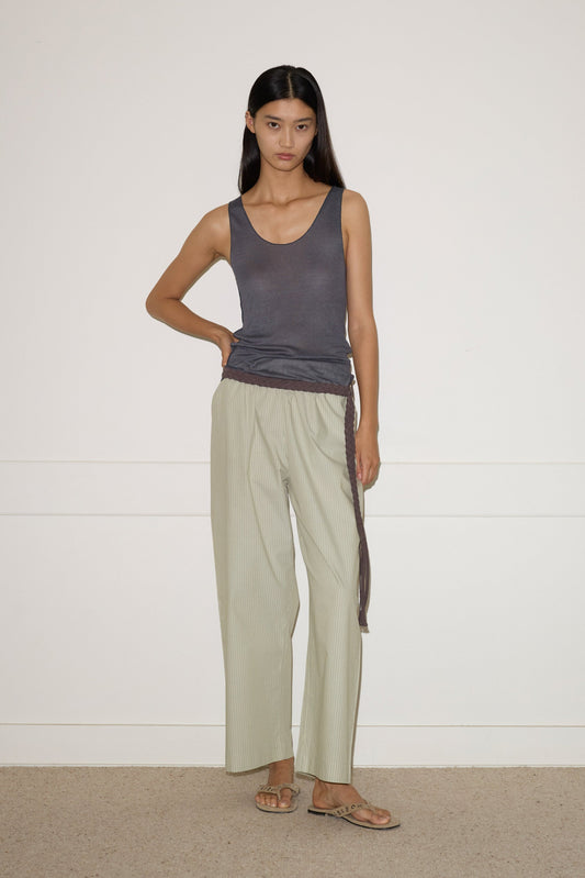 A woman with long dark hair stands against a white wall, wearing the 0 Knit Tank - Steel, light loose-fitting pants, and sandals. One hand rests on her hip while the other arm hangs by her side.