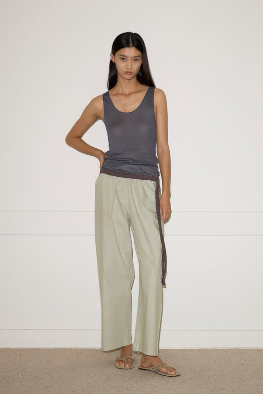 A woman stands against a plain white wall, wearing the light striped Ease Trouser, a dark gray sleeveless top, and tan sandals. She has straight dark hair and a neutral expression, with one hand resting on her hip.