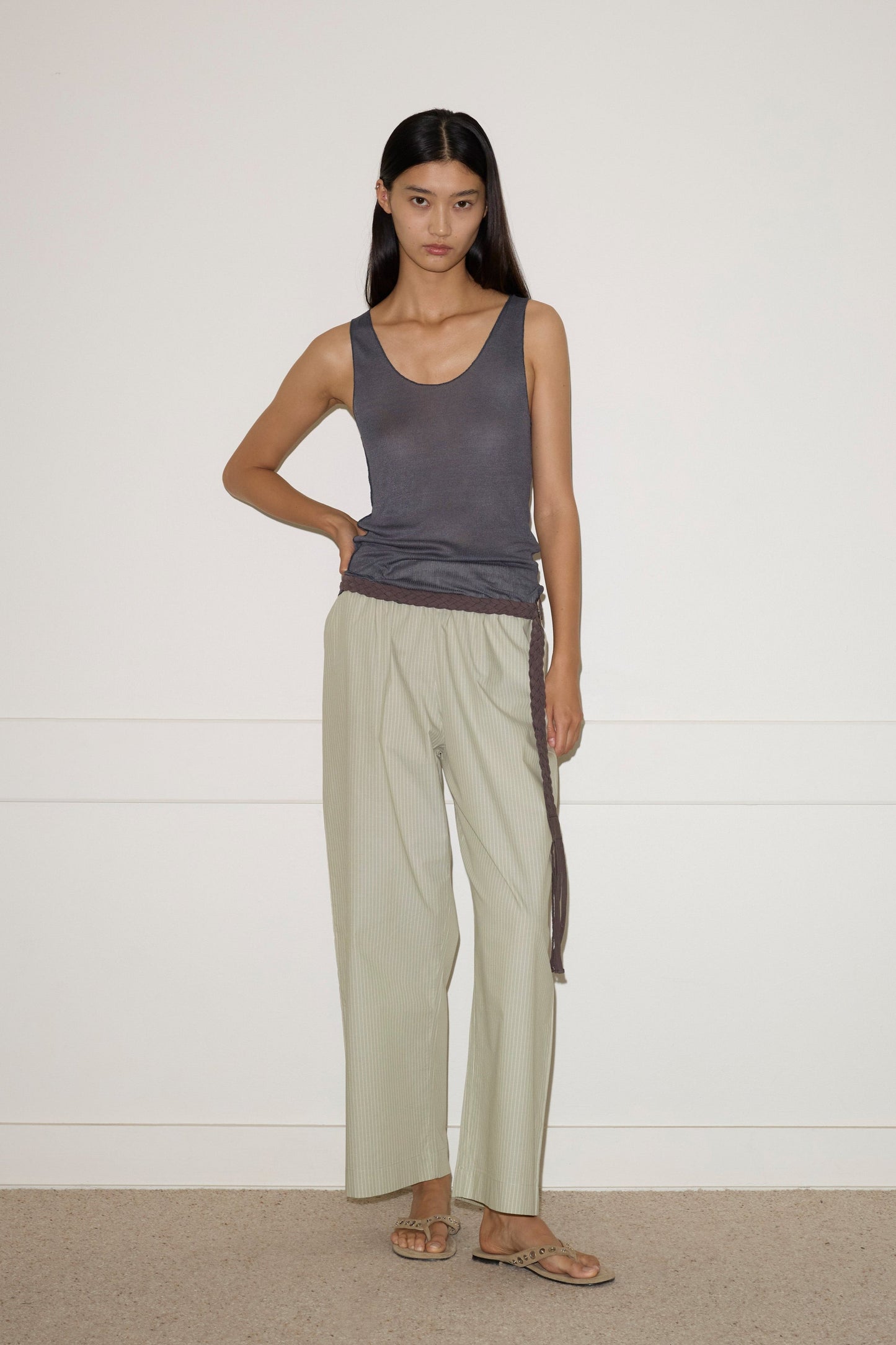 A woman stands against a plain white wall, wearing the light striped Ease Trouser, a dark gray sleeveless top, and tan sandals. She has straight dark hair and a neutral expression, with one hand resting on her hip.