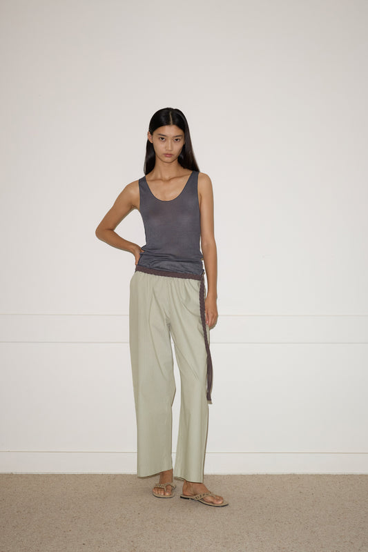 A woman with straight dark hair stands indoors against a plain white wall, wearing a sleeveless dark gray top, loose light beige organic cotton pants secured with the 0 Plaited Belt in Chocolate, and sandals. One hand rests on her hip while the other hangs by her side.